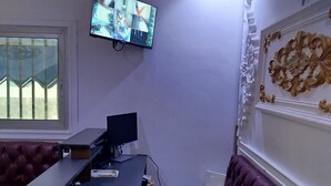 A room with a TV mounted on the wall displaying a security feed, a desk with a computer, a red leather couch, and a patterned rug. - Hôtel Famigni (Douala)