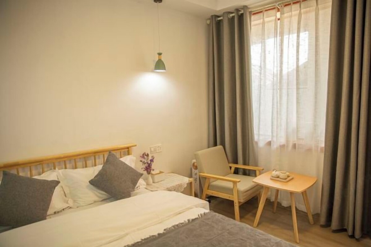 A bedroom with a bed, a chair, a small table, a lamp, and a window with curtains.