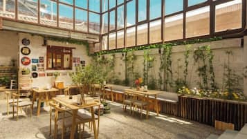 An indoor dining area with wooden tables and chairs, a high ceiling with large windows, and a wall adorned with plants.