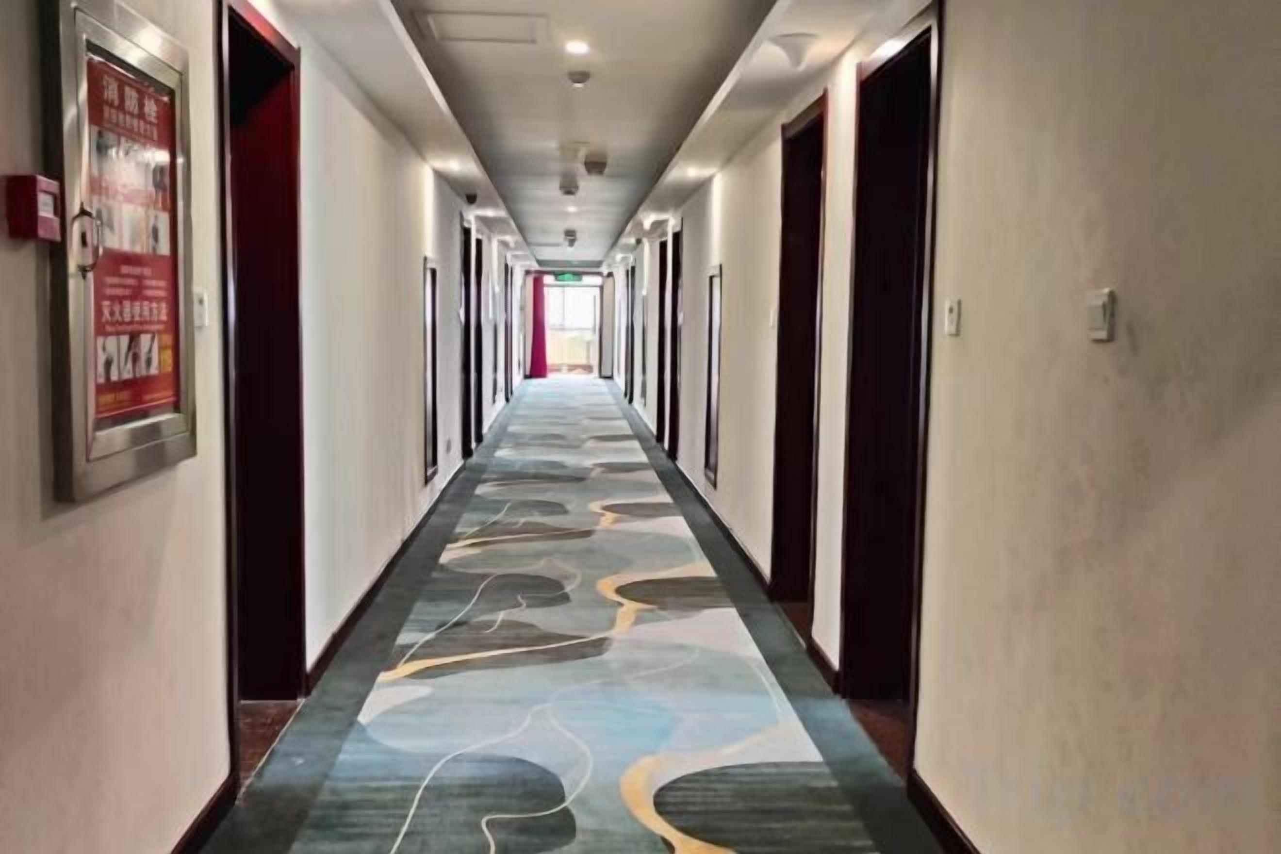 A hotel corridor with a patterned carpet, doors on both sides, and a poster on the left wall.