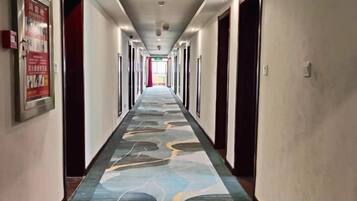 A hotel corridor with a patterned carpet, doors on both sides, and a poster on the left wall.