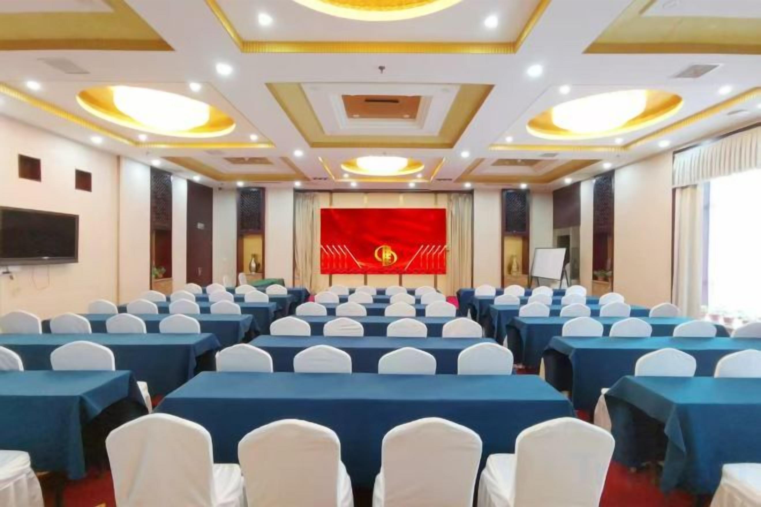 A conference room with rows of tables and chairs, a stage with a red backdrop, and a large screen.