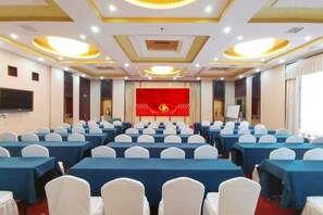 Une salle de conférence avec des rangées de tables et de chaises, une scène dotée d’un fond rouge et un grand écran.