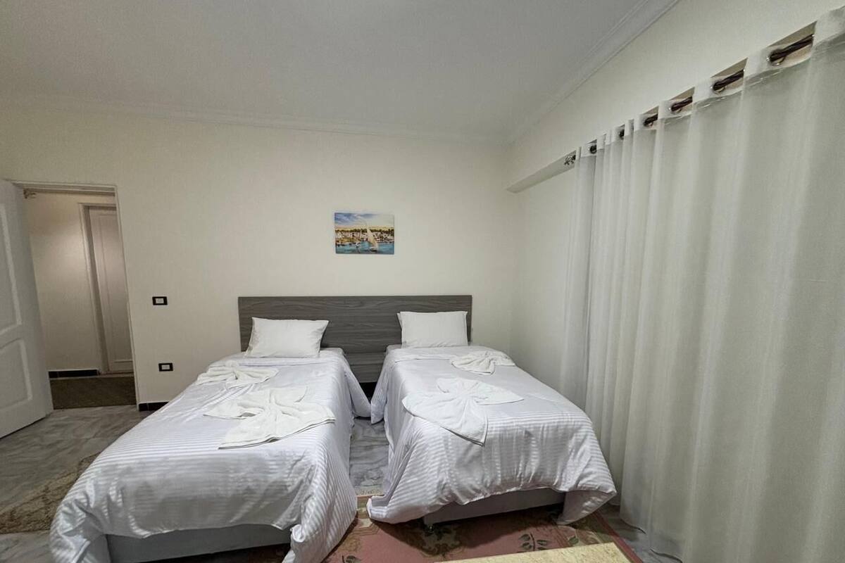 A hotel room with two beds, a headboard, a painting on the wall, and a window with curtains.
