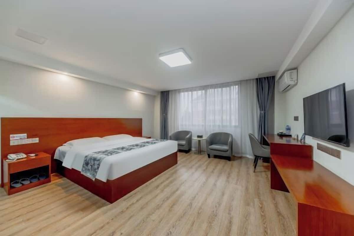Habitació d'hotel amb un llit de fusta, dues butaques, un escriptori, un televisor i una finestra amb cortines.