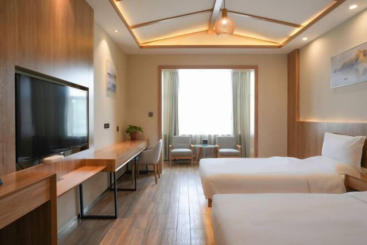 A hotel room with a large flat-screen TV, a desk with chairs, two beds, and a large window with curtains.