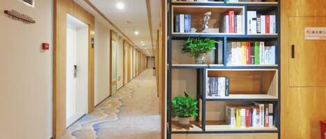 A hotel corridor with a carpeted floor, a bookshelf with books and plants, and doors leading to rooms.