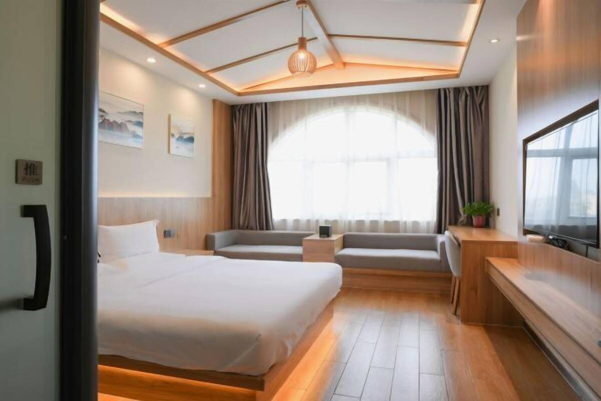 A modern hotel room with a large bed, a seating area, a TV, and a large window.
