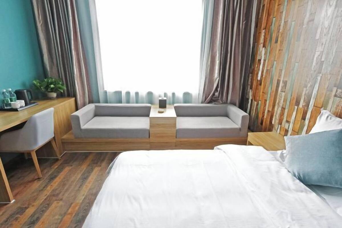 A modern hotel room with a large bed, two grey sofas, a wooden desk, and a view through a large window.