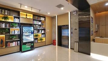 A modern hotel lobby with a vending machine, bookshelves, a reception desk, and a large mirror.