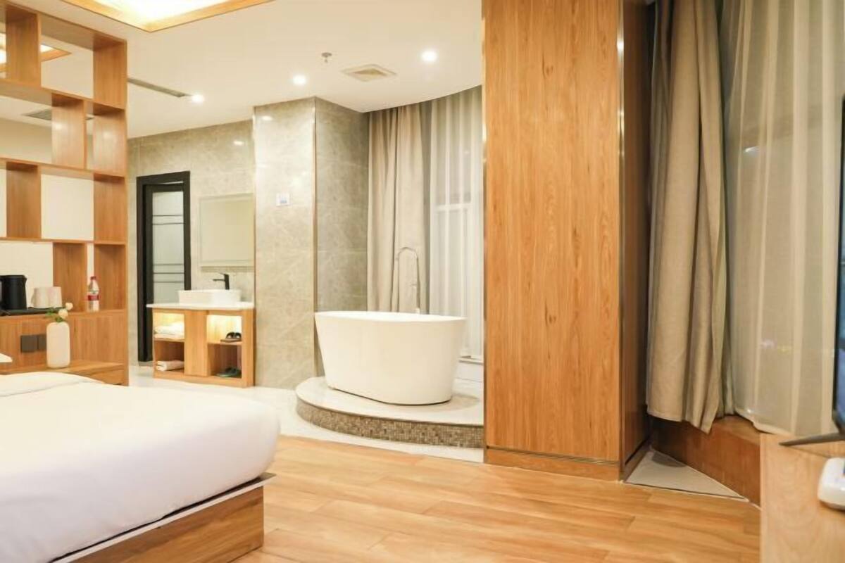 A modern hotel room with a bed, a bathroom with a bathtub, wooden shelving, and a mirror.