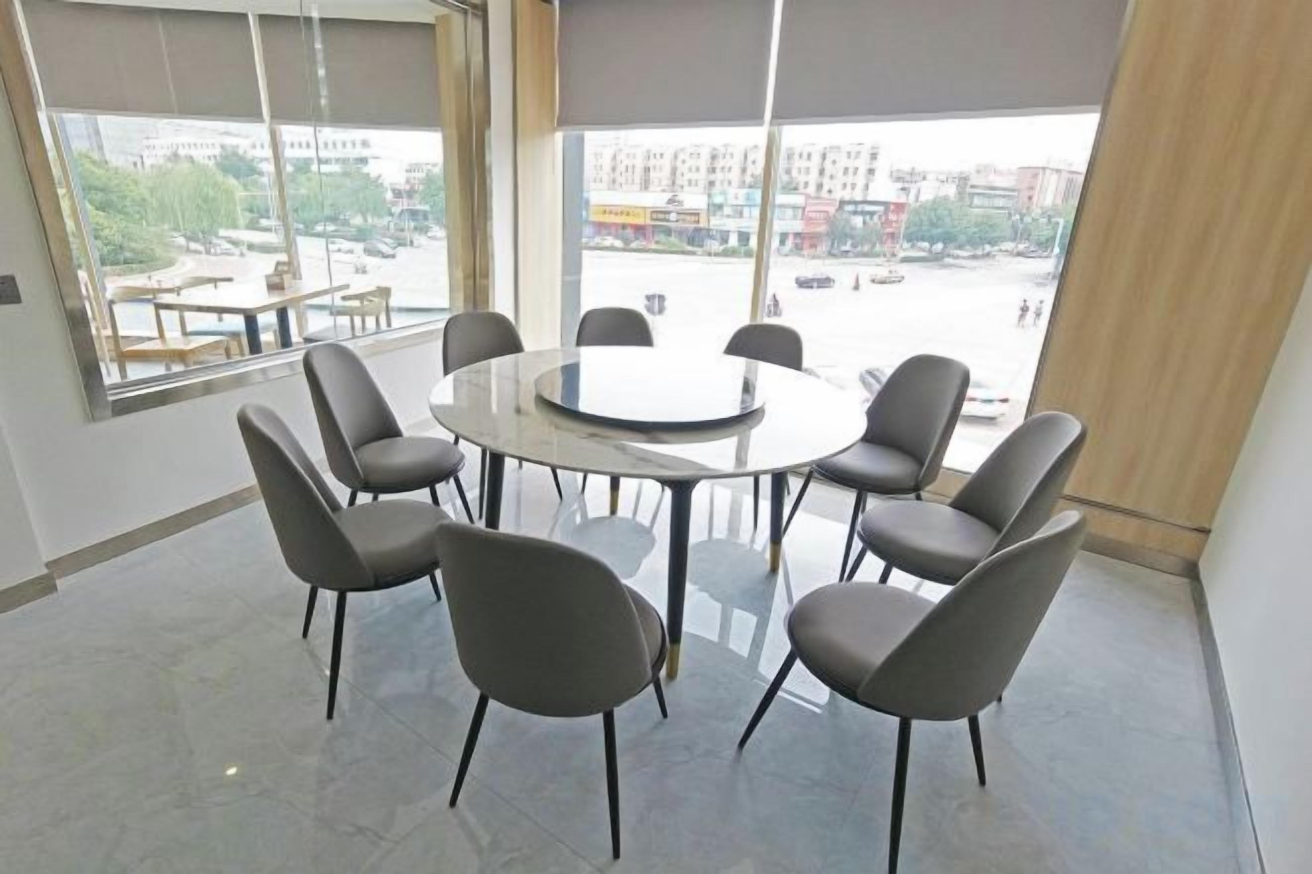 A conference room with a round table and eight chairs, large windows with blinds, and a view of the cityscape.