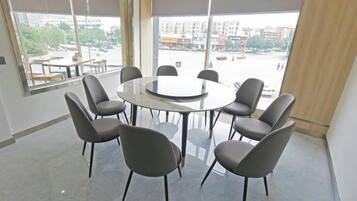 Un salón de conferencias con una mesa redonda y ocho sillas, ventanas amplias con persianas y vistas a la ciudad.