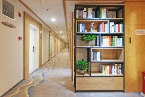 A hallway with a carpeted floor, a bookshelf with books and plants, and wooden doors on the left. - Lizun Select Hotel (Hebi Liyang Road Branch) (Hebi)
