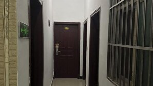 Un pasillo estrecho con una puerta de madera oscura al final, una ventana con barrotes a la derecha y un letrero direccional verde a la izquierda.