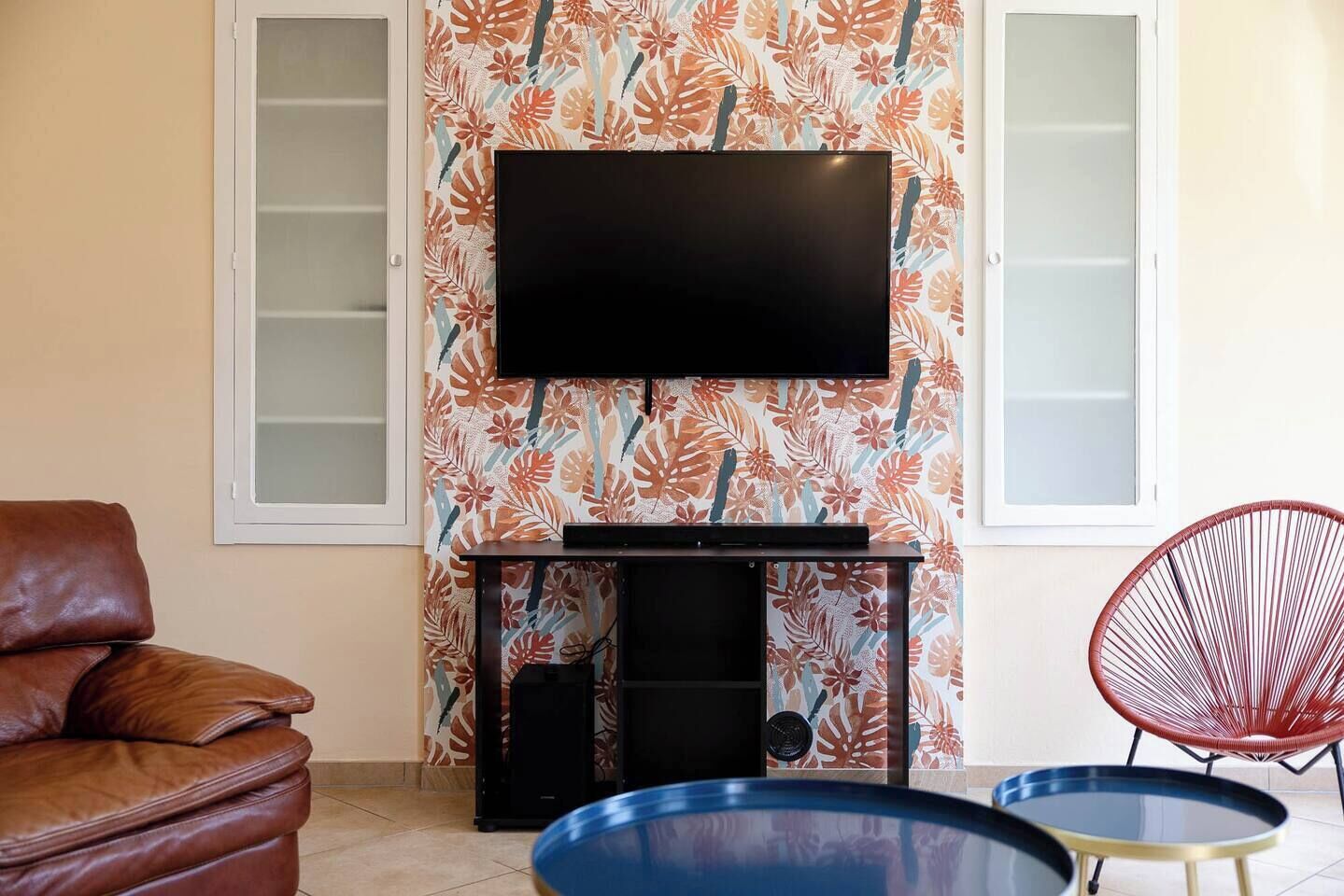 A living room with a flat-screen TV mounted on a wall with a tropical leaf pattern, a brown leather sofa, a red chair, and a small round table with a blue top.