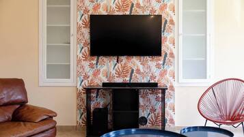 A living room with a flat-screen TV mounted on a wall with a tropical leaf pattern, a brown leather sofa, a red chair, and a small round table with a blue top.