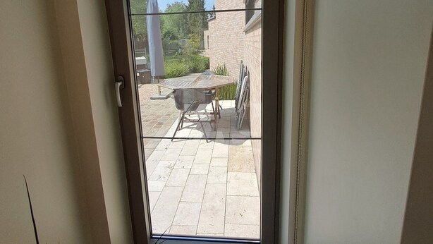 Blick durch eine Glastür auf einen Patio mit Tisch und Stühlen, einen Sonnenschirm und ein Backsteinhaus im Hintergrund.