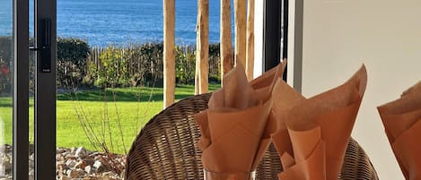 A dining table with a view of the sea, a wicker chair, and a glass table setting.