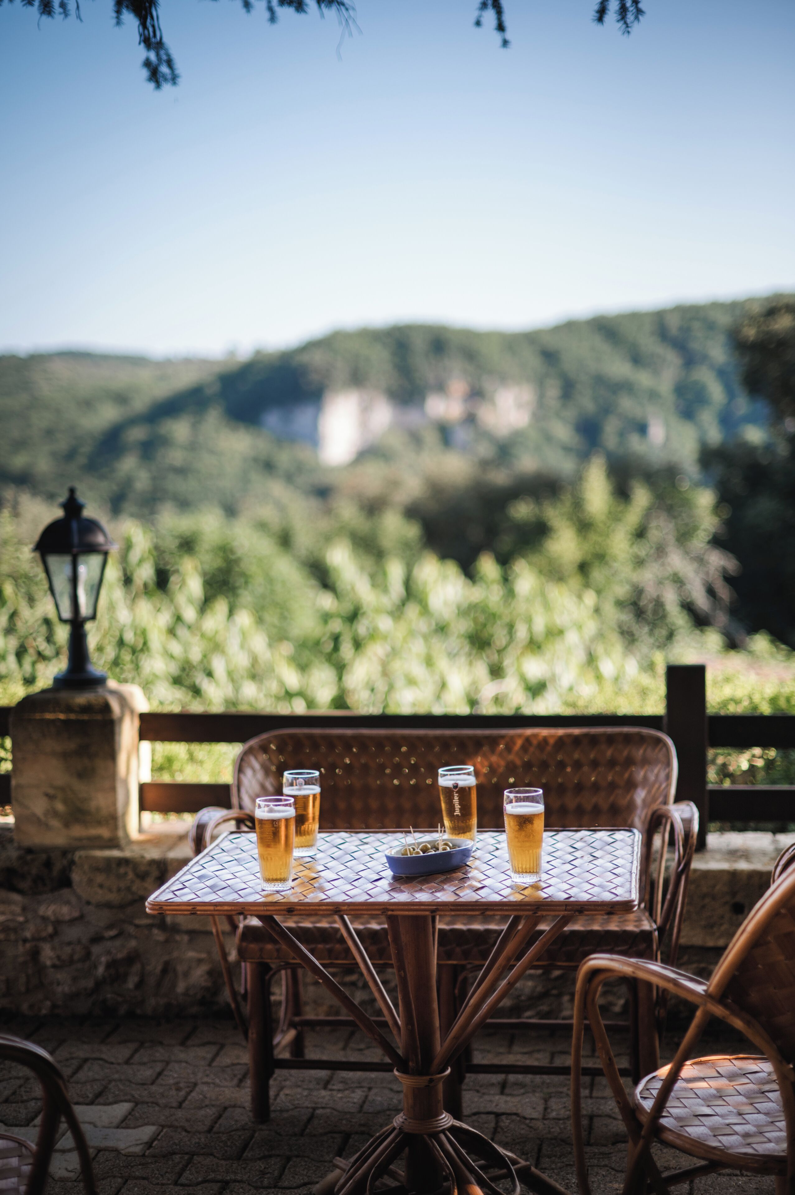 A table with four glasses of beer, a bowl of olives, and a plate, set outdoors with a view of green hills and a stone lamp post.