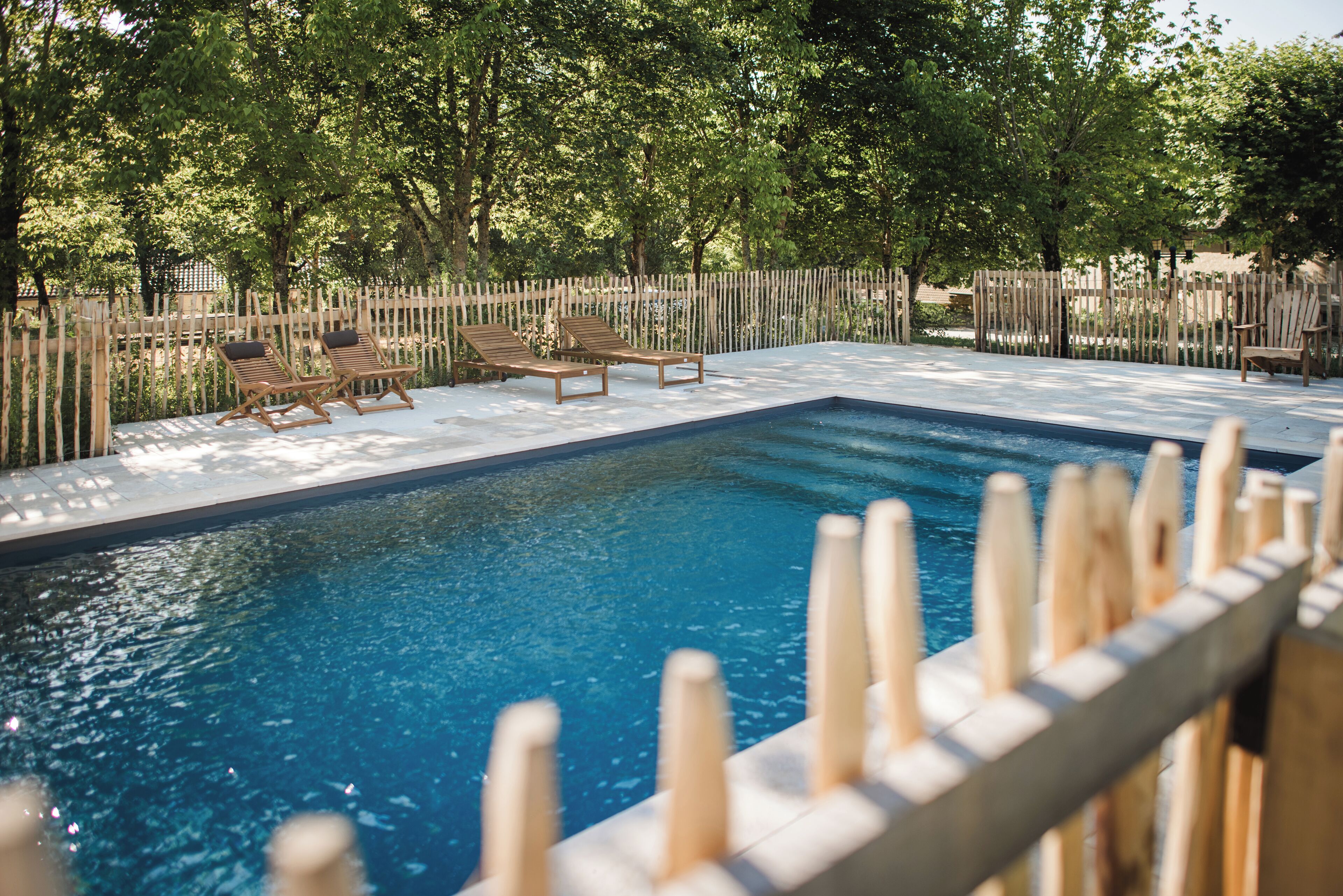 Une piscine rectangulaire entourée d’une clôture en bois, avec des chaises longues en bois disposées autour.