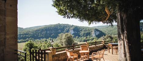 Un patio al aire libre con mobiliario de mimbre, barandillas de piedra y vistas a una ladera boscosa.