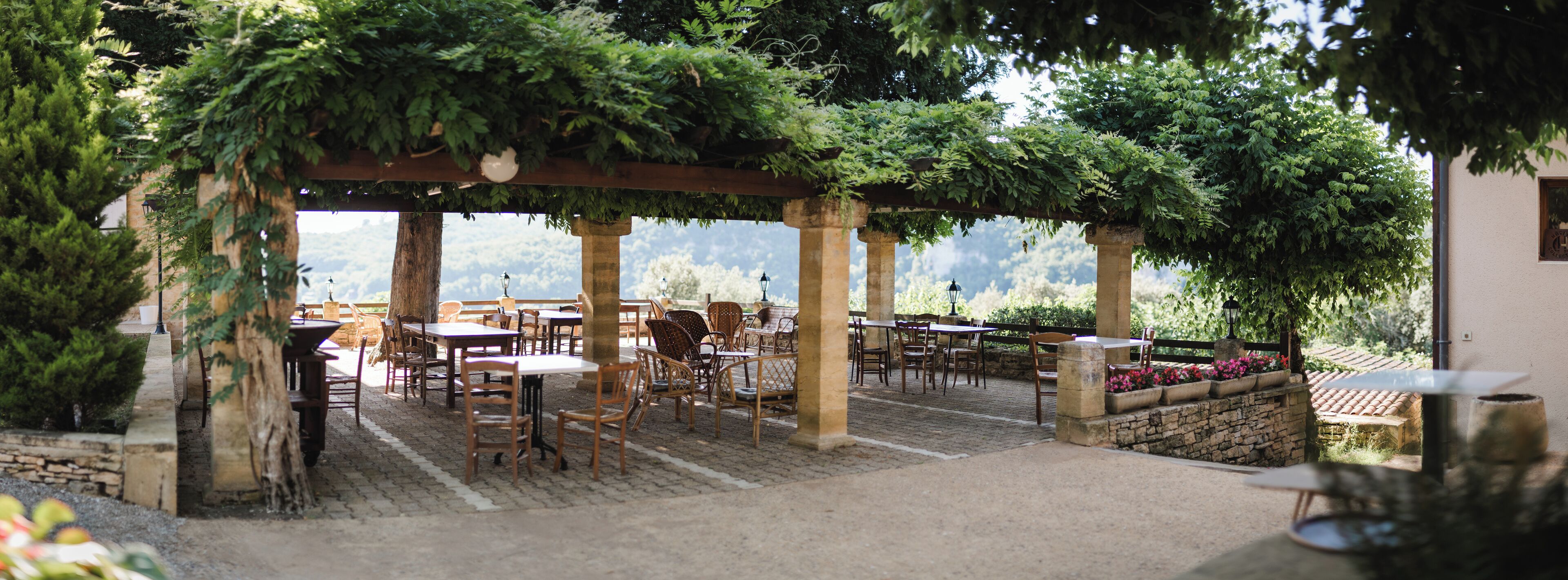 An outdoor patio with stone flooring, wooden tables and chairs, and a pergola covered with greenery.