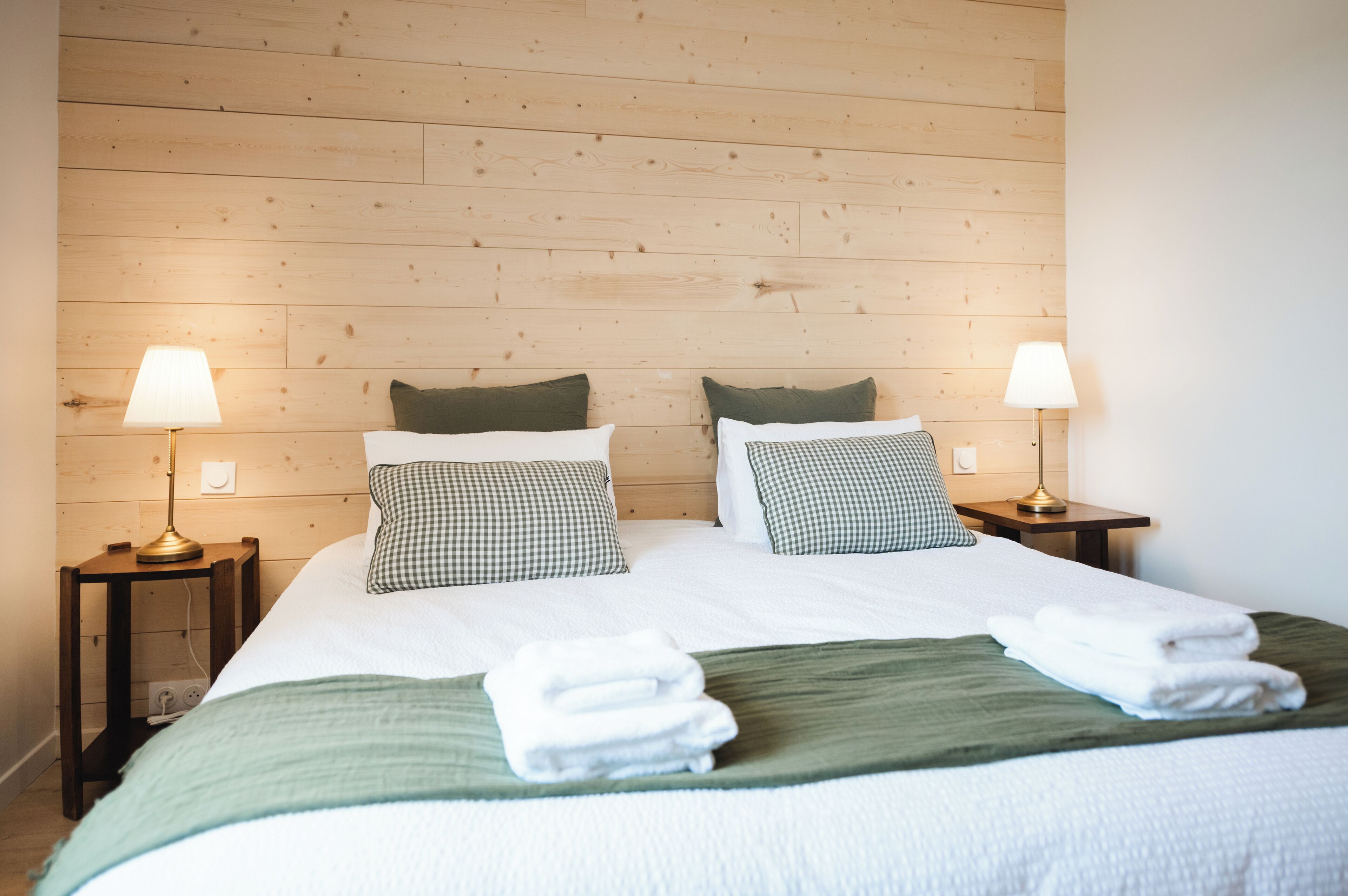Una cama bien hecha con almohadillas estampadas, una colcha verde y toallas dobladas. Hay dos mesitas de noche con lámparas y un cabecero de madera.