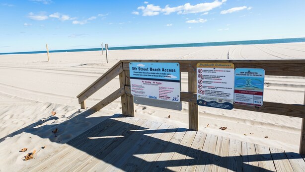 Une promenade en bois mène à une plage, et des panneaux indiquent les règles et les consignes d’accès à la plage.