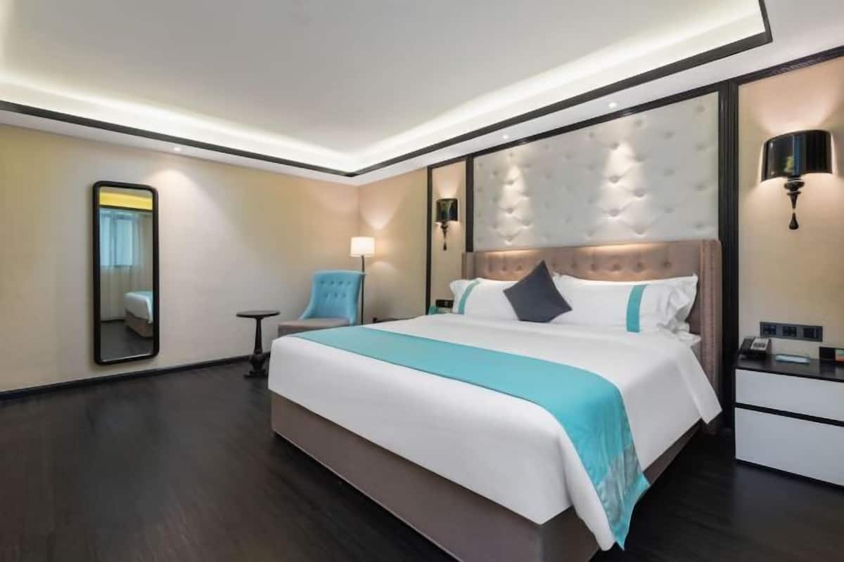 A modern hotel room with a large bed, a small side table, a chair, a mirror, and a nightstand with a lamp.
