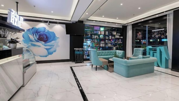 Un interior moderno con una recepción, un mural grande de rosa azul, un área de descanso con sofás turquesa y una estantería llena de libros.