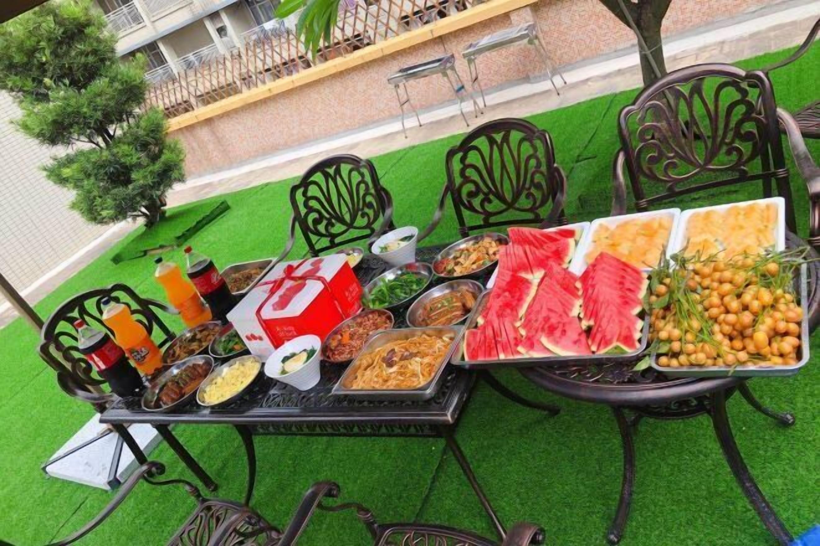 Mesa de comedor al aire libre con diversos platos, bebidas y una sandía como centro de mesa.