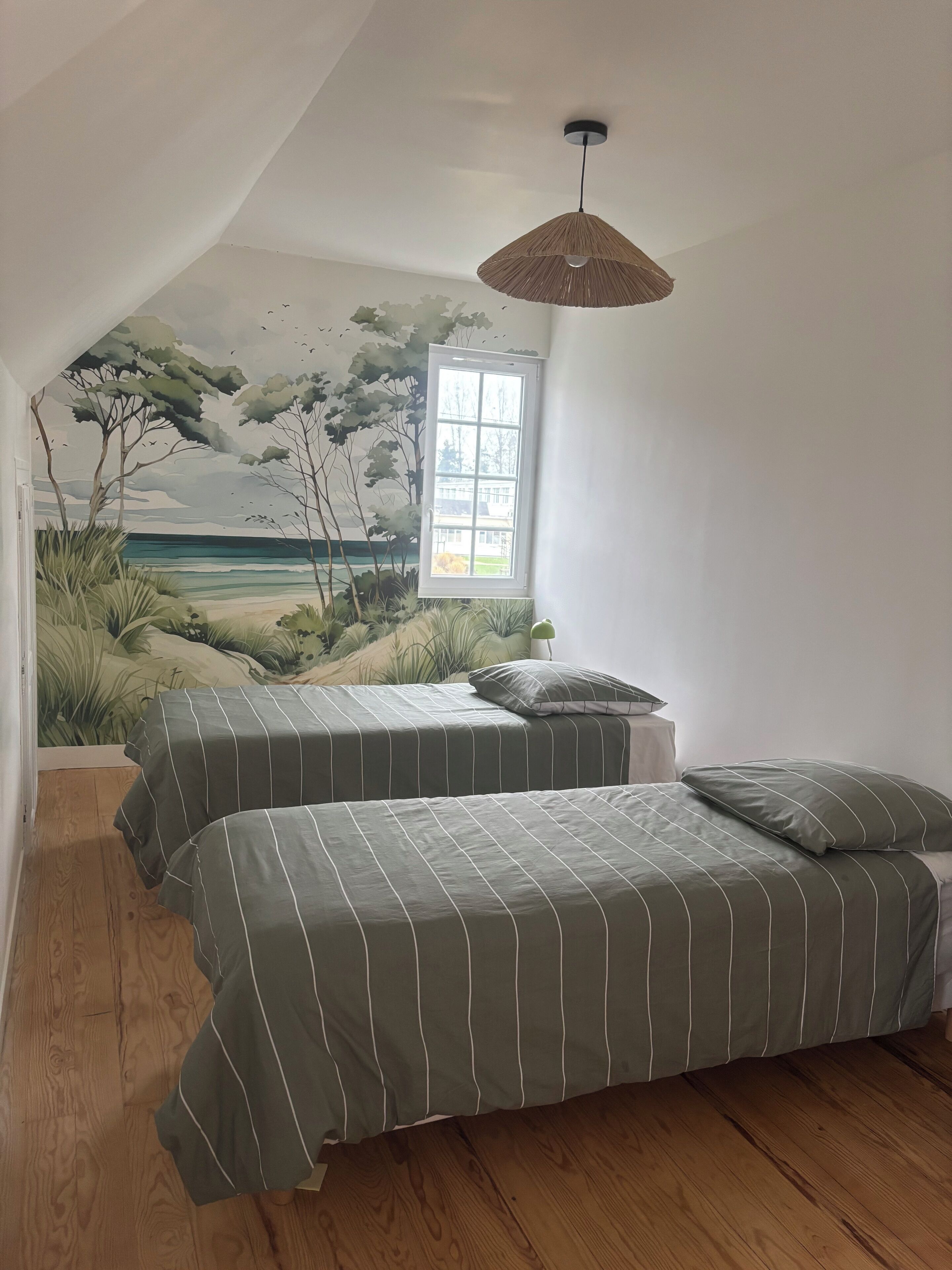 A bedroom with two beds, a mural of a beach scene, a window, and a hanging light fixture.