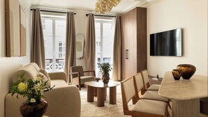 A modern living room with a sofa, chairs, a coffee table, and a dining table. There is a large window with curtains, a flat-screen TV mounted on the wall, and a decorative vase with flowers on the coffee table. - Elegant Apartment - 2br- 6P- Gare Saint Lazare (Paris)