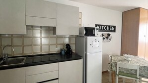 Une cuisine compacte avec des meubles blancs, un plan de travail noir, un four à micro-ondes et un réfrigérateur. On y trouve une petite table à manger recouverte d’une nappe à motifs et une plante en pot posée sur le sol.