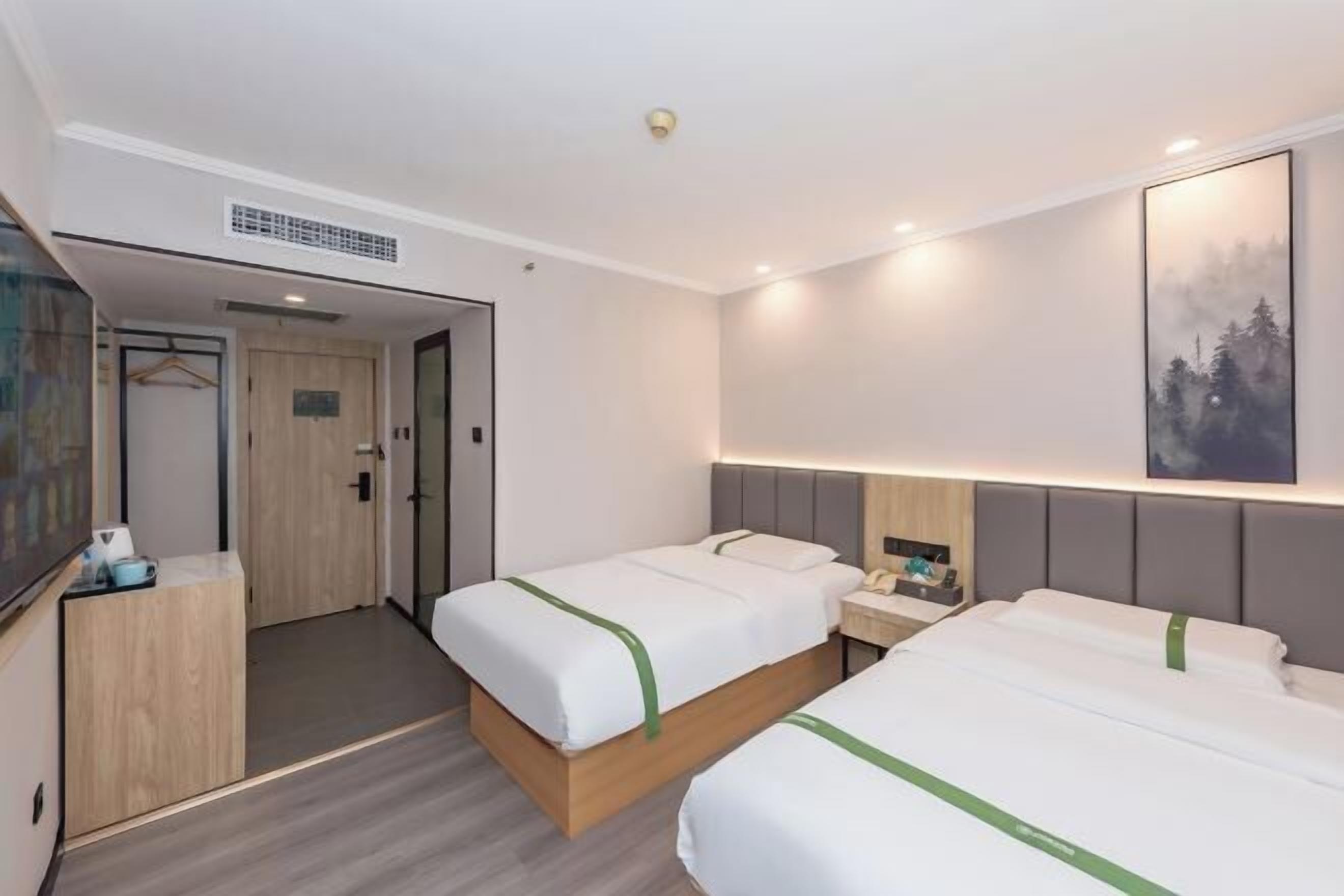 A hotel room with two beds, a bedside table with a lamp, a painting on the wall, and a door leading to another room.