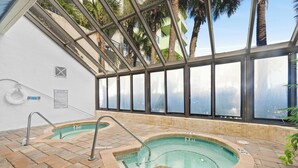 A glass-enclosed indoor pool with two hot tubs, surrounded by a tiled deck and a view of palm trees and buildings. - Coral Beach 1022-Oceanfront 1BR-Pools-Lazy River (Myrtle Beach)