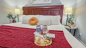 A hotel room with a neatly made bed, a red throw blanket, a tray with bottled water and snacks, and a wall art piece.