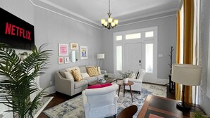 A living room with a sofa, armchair, coffee table, and a dining table with chairs. There is a plant, a lamp, and a wall-mounted TV displaying the Netflix logo.