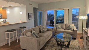 A modern living room with a sofa, two armchairs, a glass coffee table, and a kitchen area with a microwave and coffee maker.