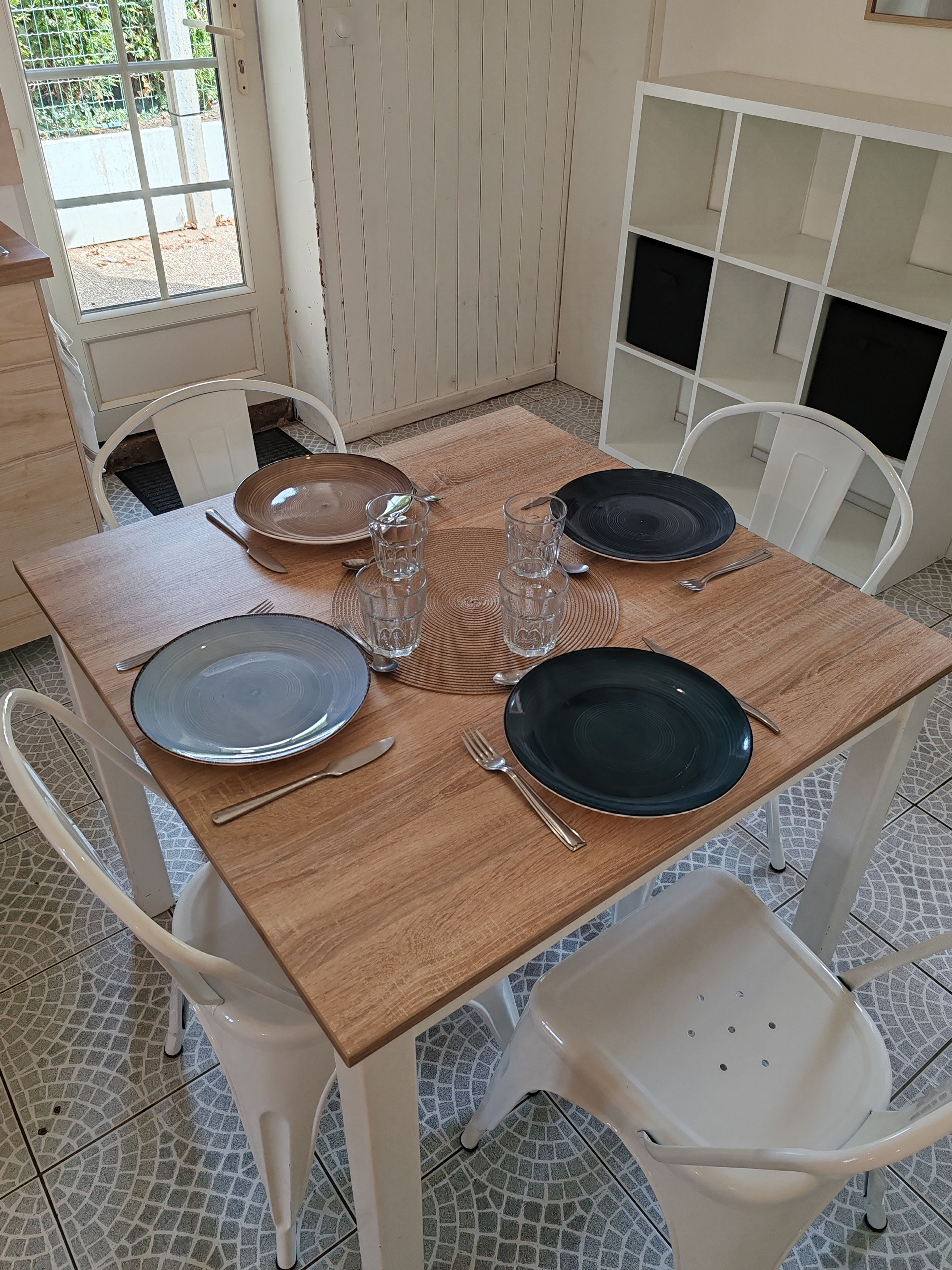 Une table à manger, dressée avec des assiettes, des couverts et des verres, entourée de chaises blanches, avec un sol carrelé et une porte donnant sur l’extérieur.
