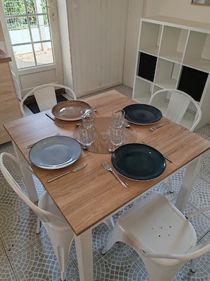 Een eettafel is gedekt met borden, bestek en glazen, omringd door witte stoelen, met een betegelde vloer en een deur naar buiten.