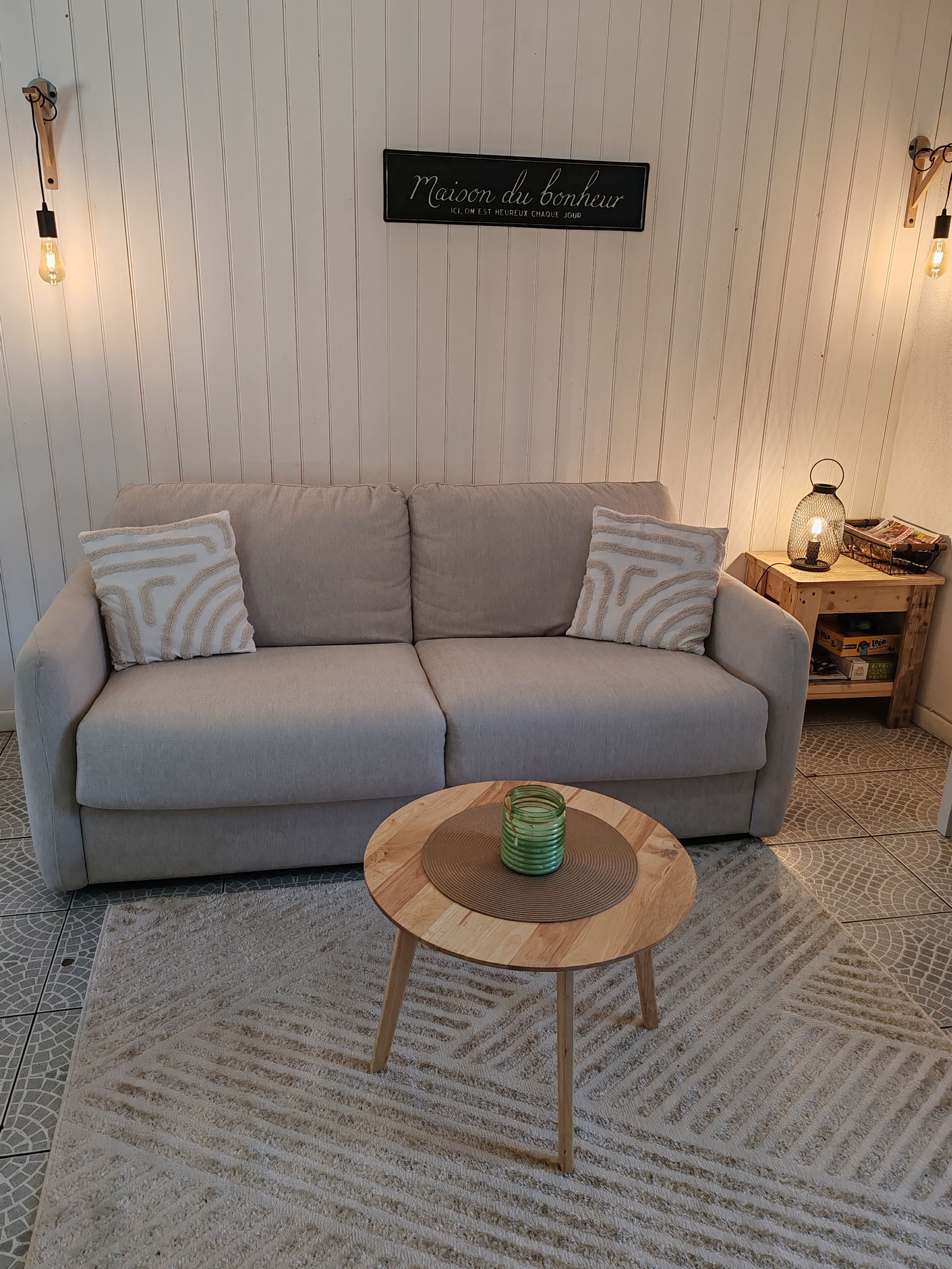 A neutral-toned sofa with patterned cushions, a round wooden coffee table with a glass jar, and a wall-mounted sign.