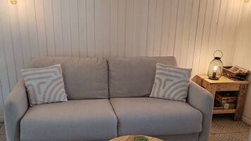 A neutral-toned sofa with patterned cushions, a round wooden coffee table with a glass jar, and a wall-mounted sign.