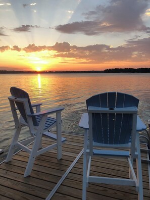 A wooden deck with two Adirondack chairs facing a sunset over a calm body of water.
