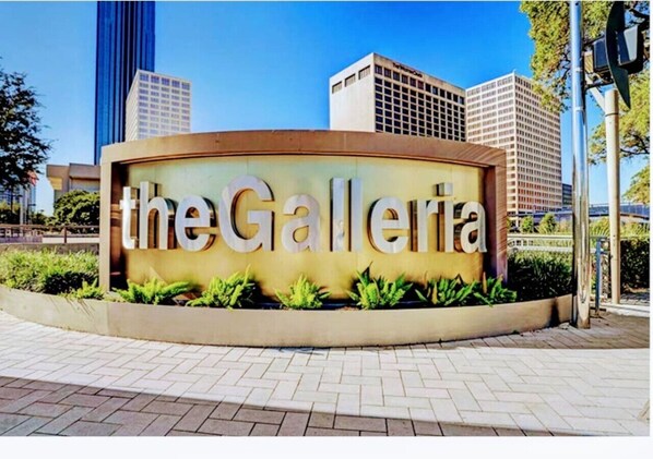 Señal de 'the Galleria' con un diseño moderno, rodeada de áreas ajardinadas y edificios urbanos al fondo.