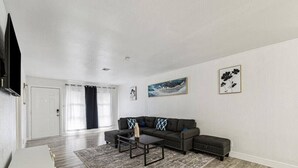 A spacious living room with a dark-colored sectional sofa, a coffee table, and a patterned rug. There are two framed artworks on the walls and a door leading to another room. - Serene 3BR Luxe Easy Access to NRG & Galleria (Houston)