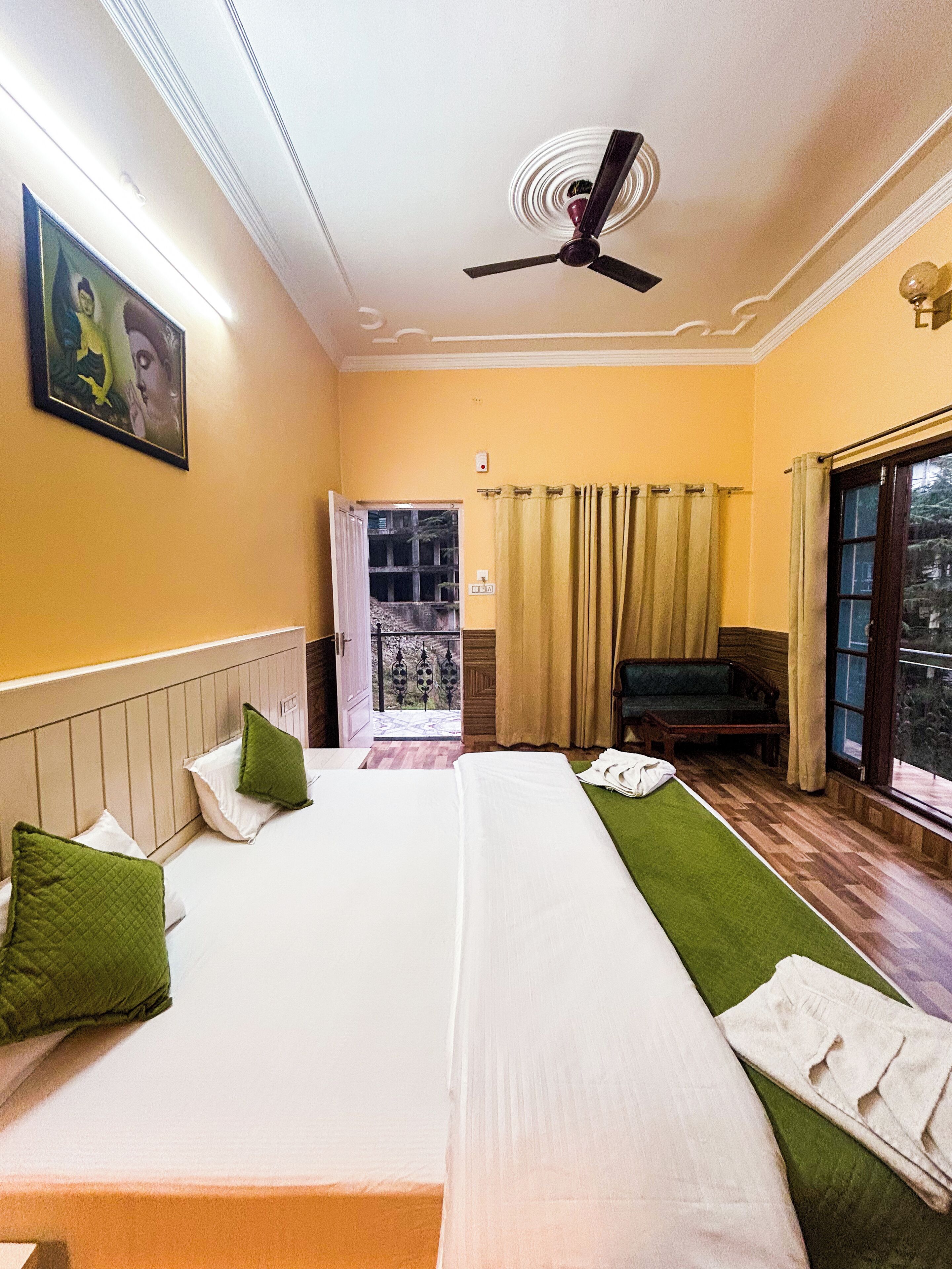 A hotel room with two beds, a ceiling fan, a painting on the wall, a window with curtains, and a view of the outside.
