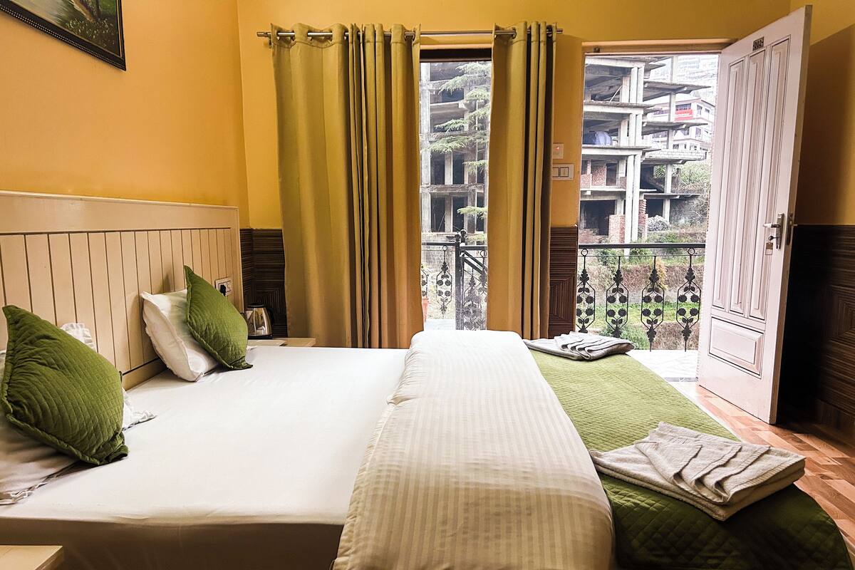 A hotel room with a bed, green pillows, a window with curtains, and a view of buildings outside.