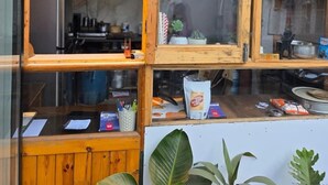 Una finestra con cornice in legno che si affaccia su un panorama urbano. All'interno, si vedono scaffali con diversi oggetti e un bancone con un sacchetto di patatine.